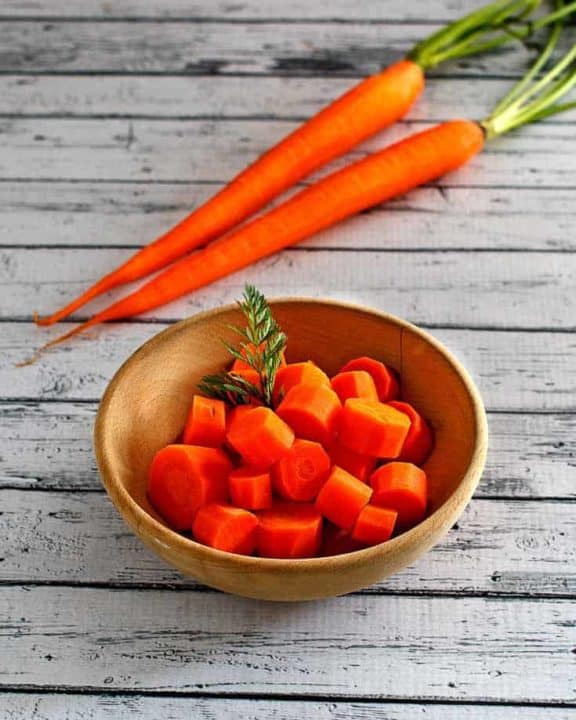 a bowl of cooked carrots next to two fresh carrots