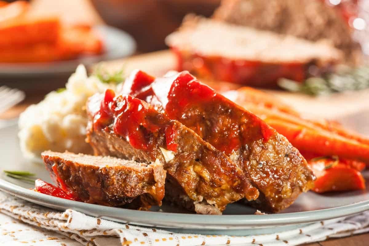 Sauce topped meatloaf slices on a plate with carrots and mashed potatoes.
