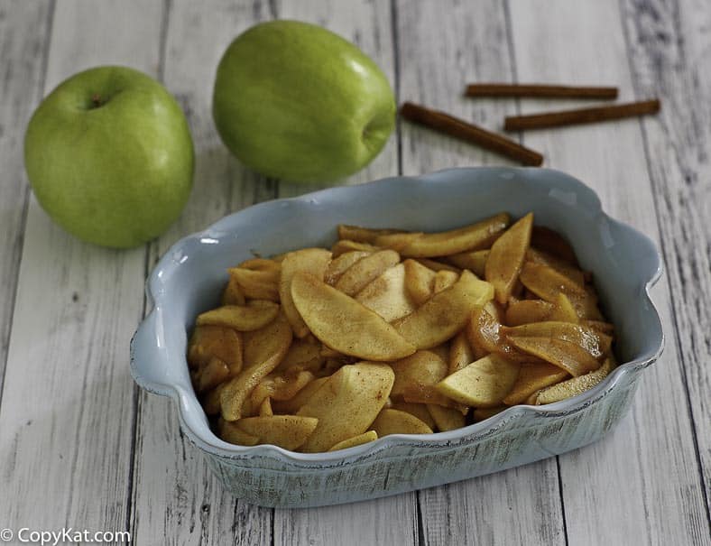 Homemade Cracker Barrels fried apples in a serving dish next to 2 apples and cinnamon sticks.