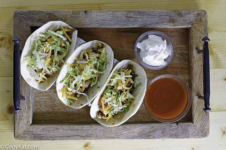 A bowl of homemade Taco Bell taco sauce and 3 tacos on a wood platter.