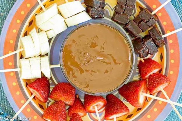 A fondue pot full of chocolate fondue with fruit skewers around it.