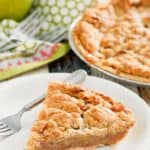 a slice of Dutch apple pie and a fork on a plate with the pie and apples in the background