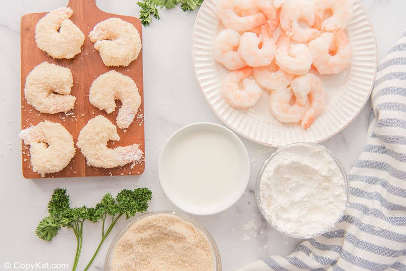 panko breaded shrimp on a wood board, raw shrimp, and breading ingredients in bowls