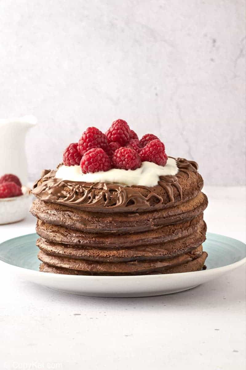 a stack of six chocolate pancakes topped with chocolate spread, whipped cream, and raspberries