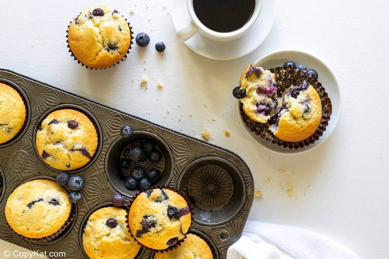 blueberry muffins in a muffin tin and on a plate and a cup of coffee