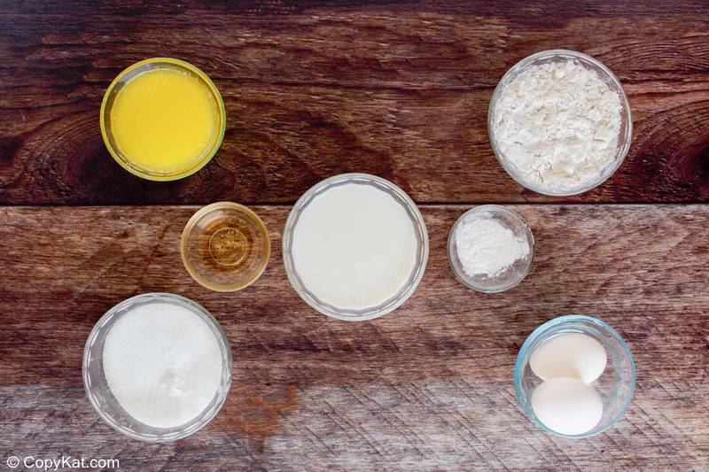 An overhead view of ingredients for almond cake in glass bowls on a wood table: sugar, eggs, milk, flour, butter, and almond extract.