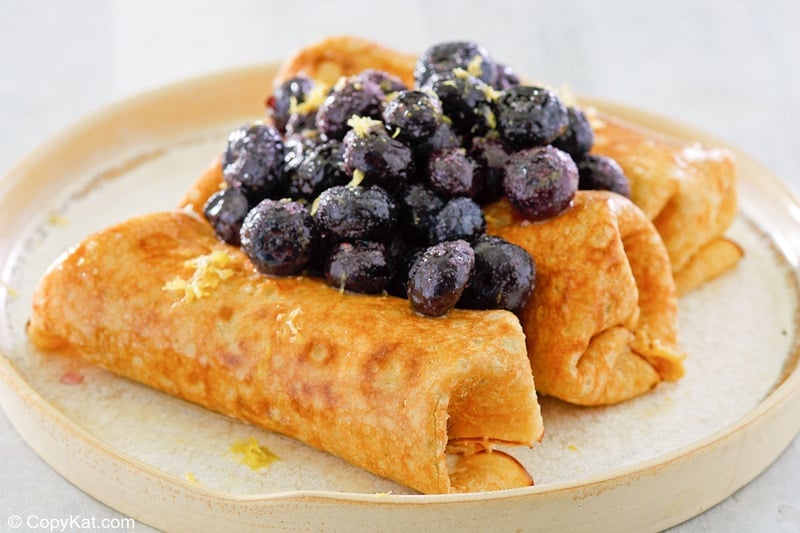 three cheese blintzes topped with blueberries and lemon zest on a plate.