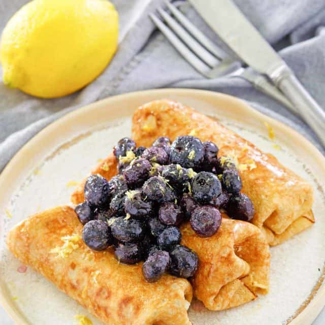 three cheese blintz topped with blueberries on a plate, a lemon, knife, and fork.