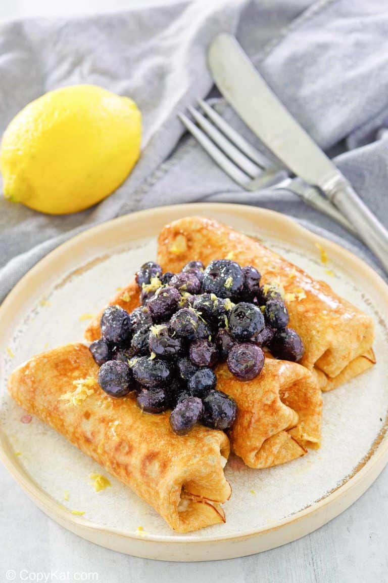 three cheese blintz topped with blueberries on a plate, a lemon, knife, and fork.
