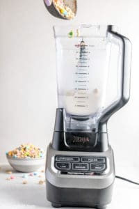 adding Lucky Charms cereal to a milkshake mixture in a blender.
