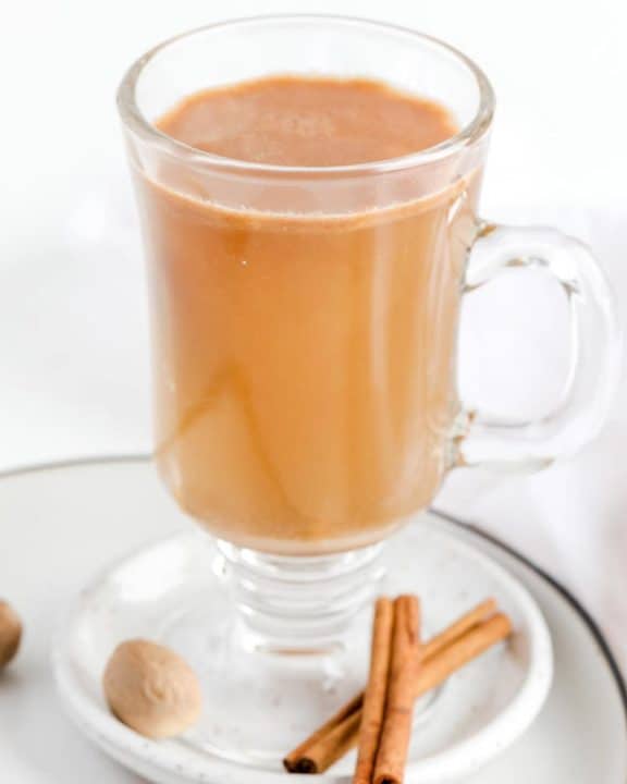 hot buttered rum in a glass mug on a plate with cinnamon sticks and nutmeg.