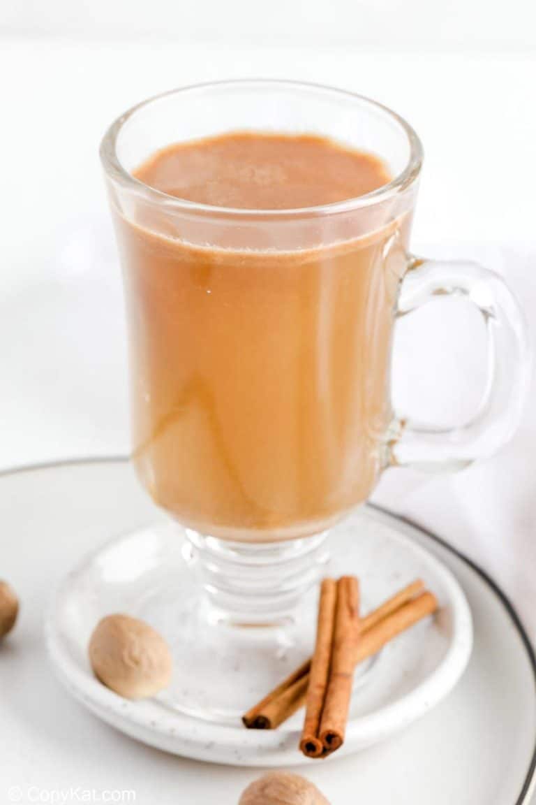 hot buttered rum in a glass mug on a plate with cinnamon sticks and nutmeg.