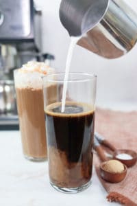 pouring frothed milk into a glass with espresso and caramel syrup.