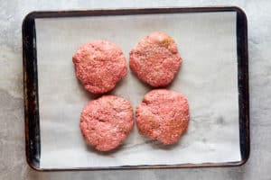 four seasoned raw hamburger steak patties on a baking sheet.