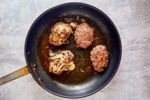 browned ground beef patties topped with mushrooms and onions in a skillet.