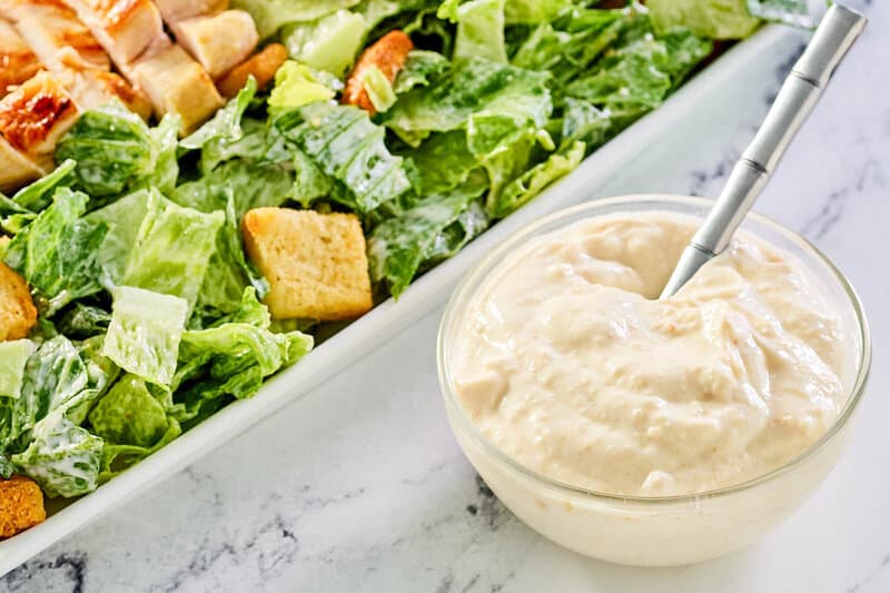 small bowl of creamy caesar dressing next to a caesar salad.