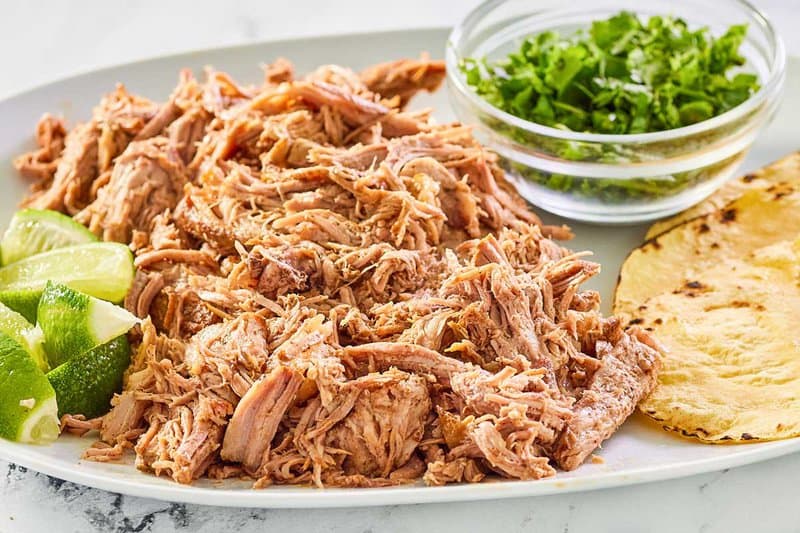 shredded pork shoulder, limes, tortillas, and fresh cilantro on a platter.