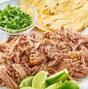 pulled oven roasted pork shoulder on a platter with limes, cilantro, and tortillas.