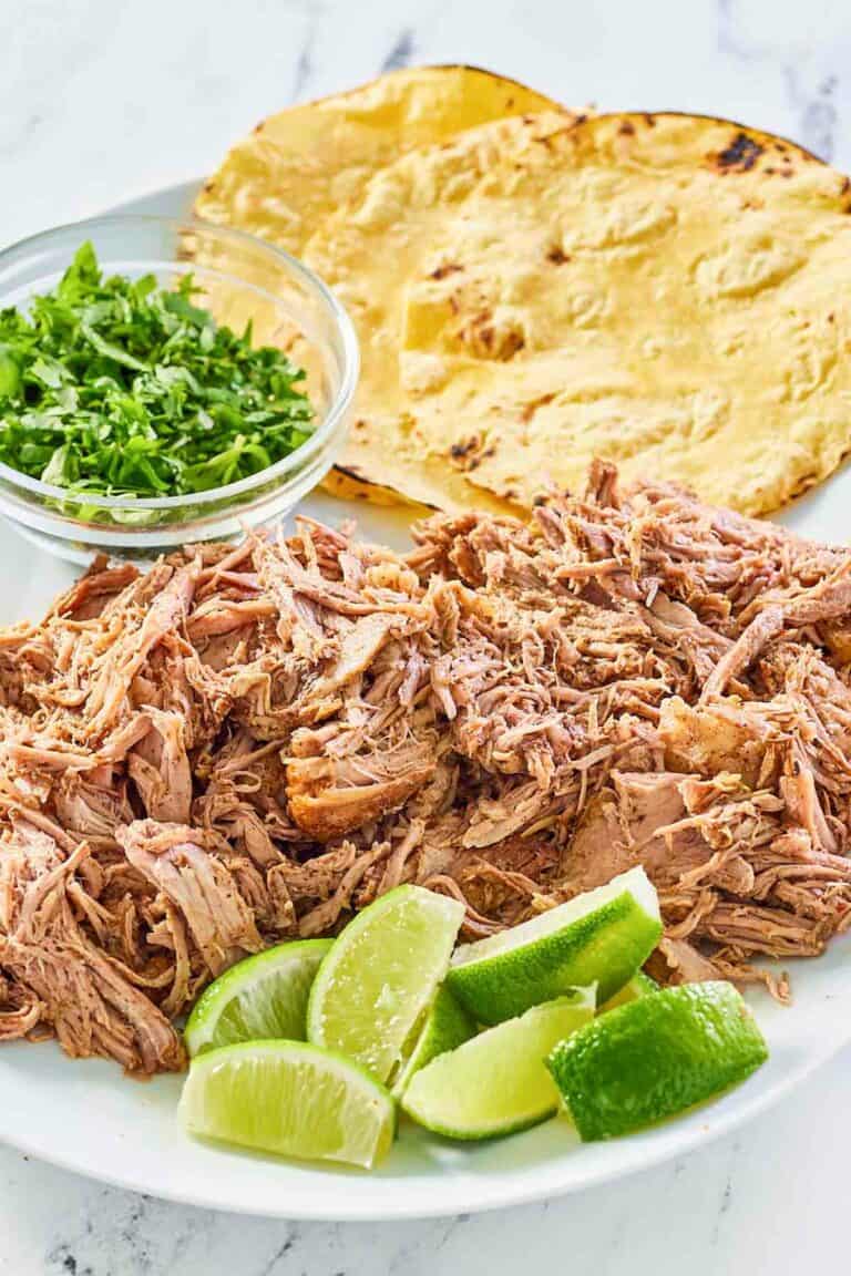 pulled oven roasted pork shoulder on a platter with limes, cilantro, and tortillas.