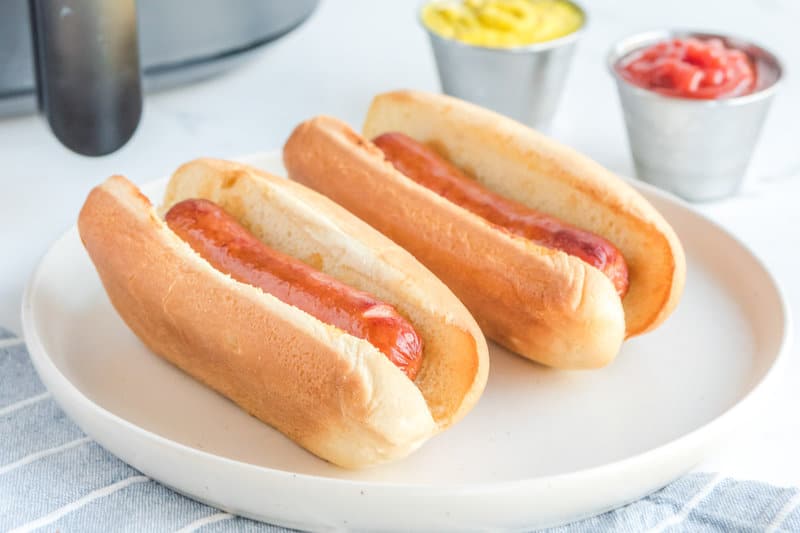 Two air fried hot dogs in buns in a plate in front of an air fryer.