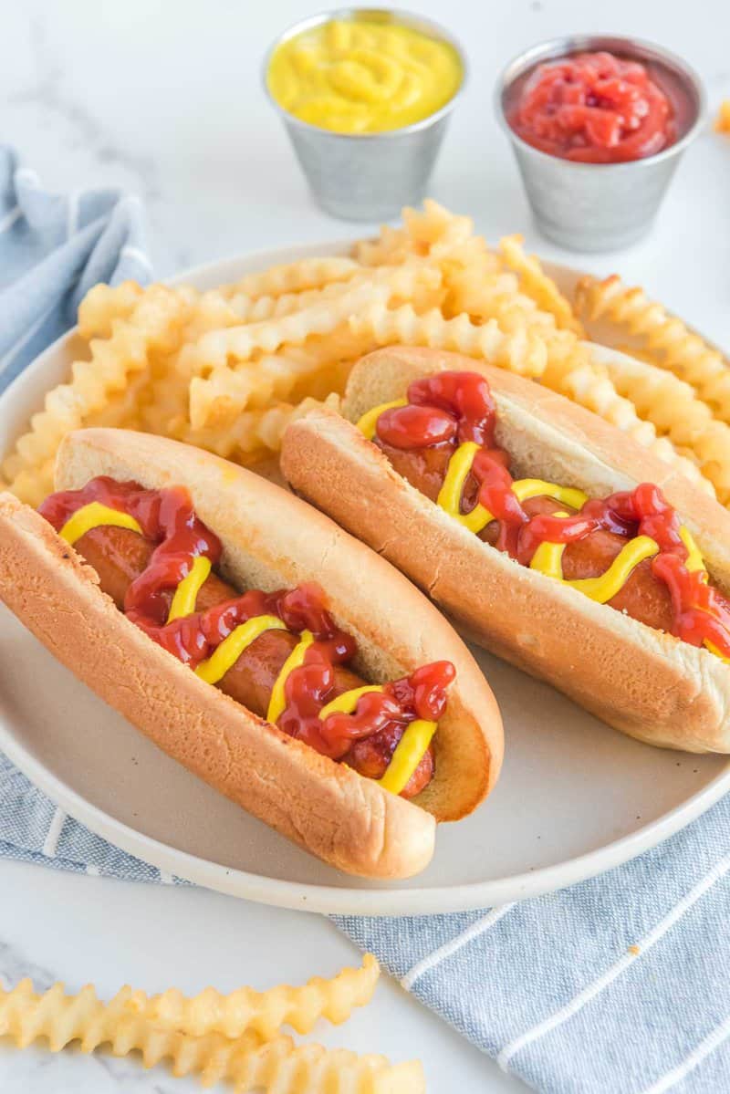 Two air fried hot dogs in buns with ketchup and mustard on a plate with fries.