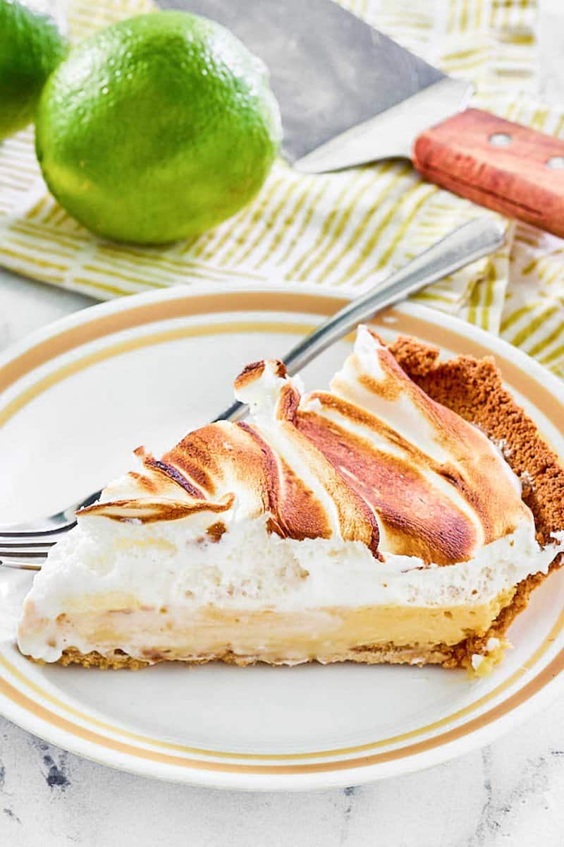 Key lime meringue pie slice on a plate, a lime, and a pie server.