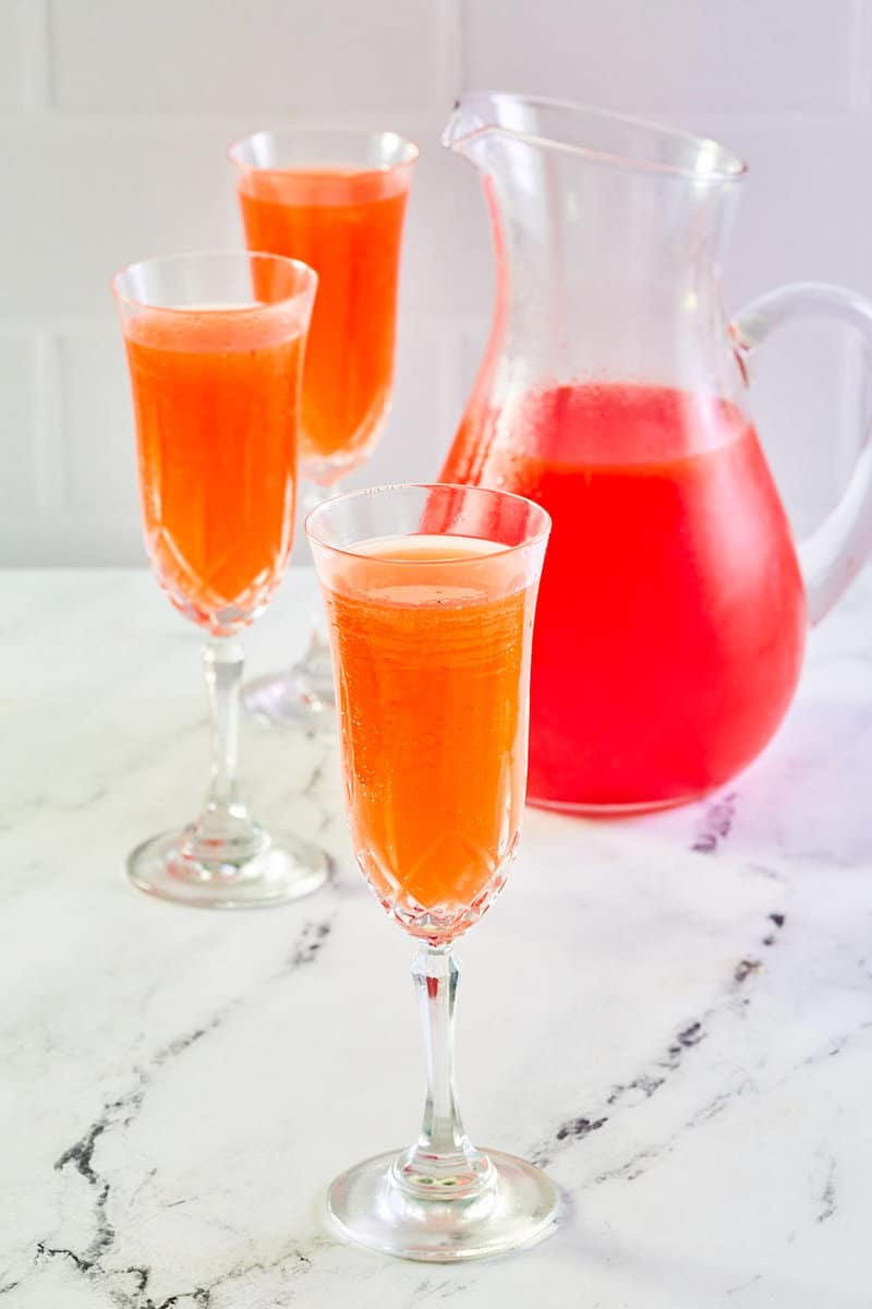 Strawberry mimosa in three champagne flutes and a pitcher.