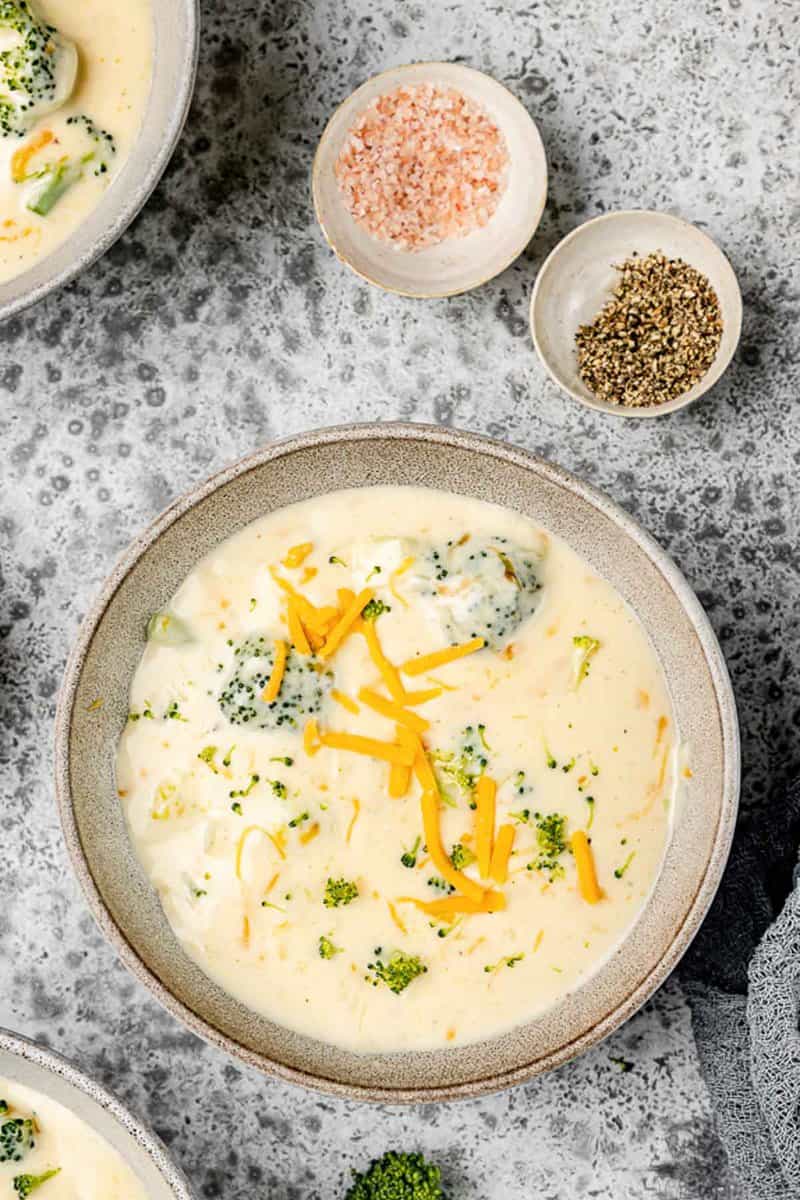 Overhead view of three cheese broccoli soup and bowls of pink salt and black pepper.