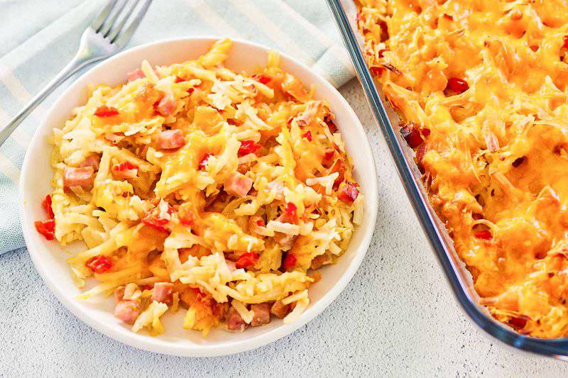 Cheesy hash brown casserole with ham on a white plate and in a glass baking dish.
