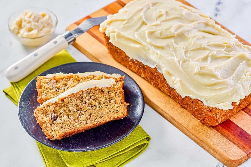 Homemade zucchini nut bread with cream cheese frosting loaf on a board and slices on a plate.