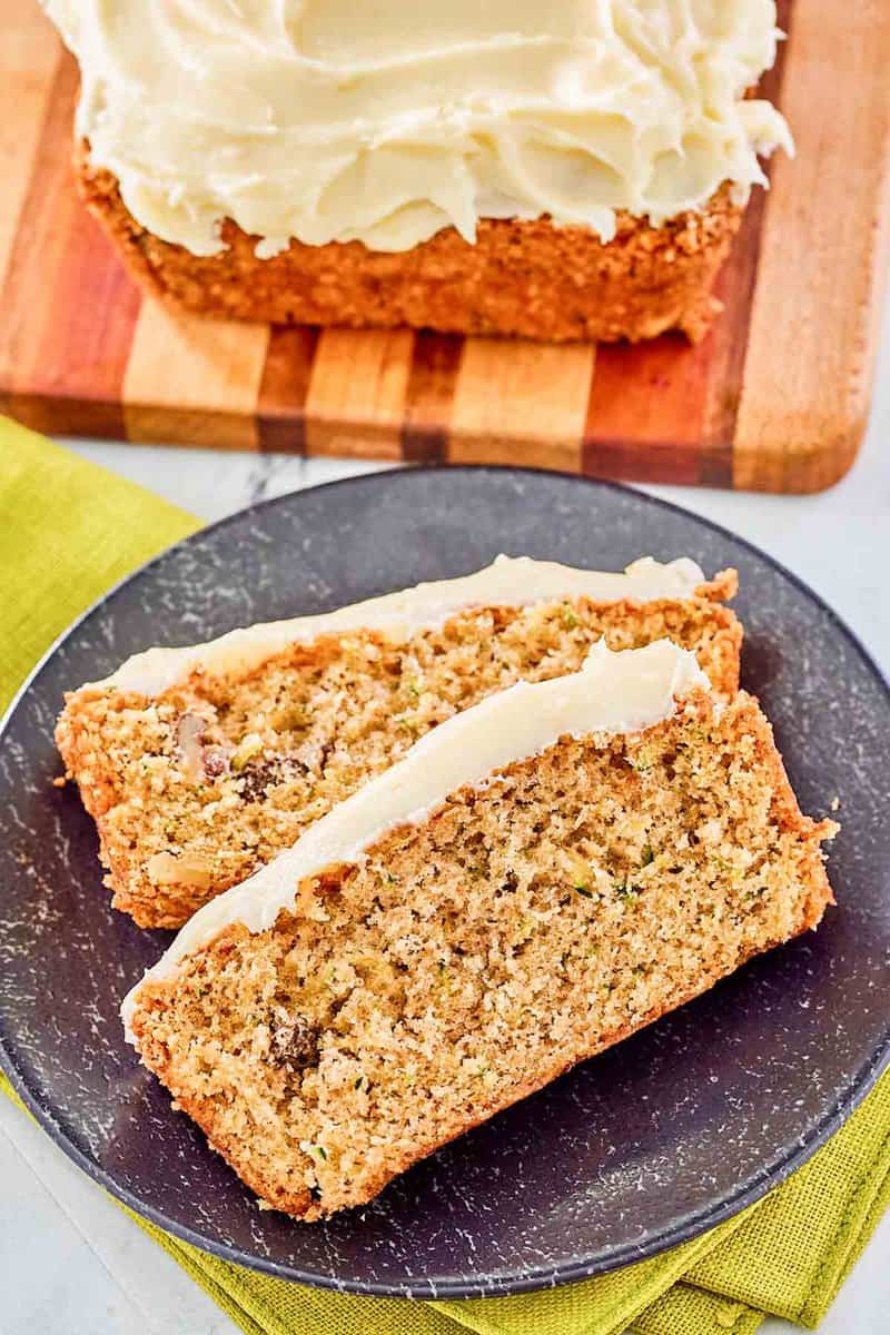 Zucchini nut bread with cream cheese frosting slices on a plate and a loaf on a wood board.
