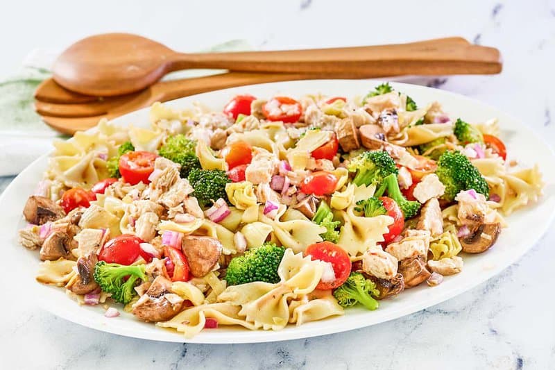 Chicken pasta salad with vegetables on a platter and salad servers behind it.