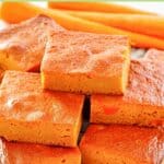 Carrot pudding slices stacked on a plate and fresh carrots behind them.