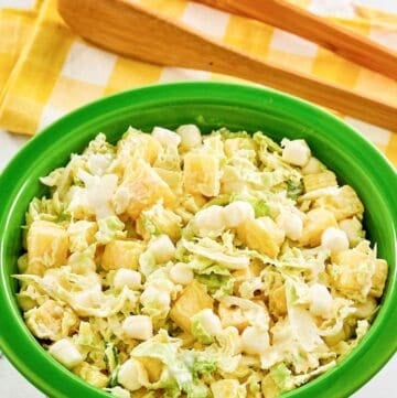 A bowl of pineapple coleslaw and salad tongs behind it.