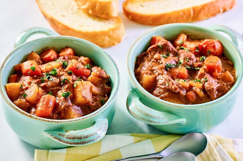 Crockpot beef stew in bowls and bread slices behind them.