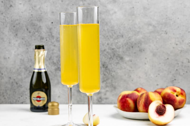 Two copycat Olive Garden peach bellini drinks, peaches, and bottle of sparkling wine.