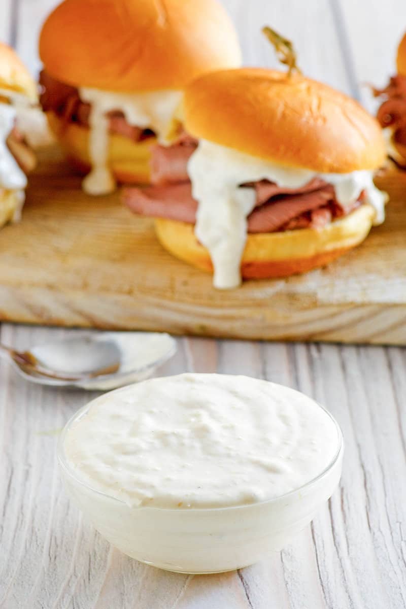 A bowl of copycat Arby's horsey sauce and roast beef sandwiches behind it.