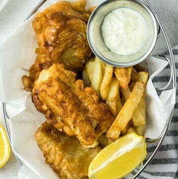 Beer battered fish and chips, lemon wedge, and a cup of tartar sauce in a serving basket.