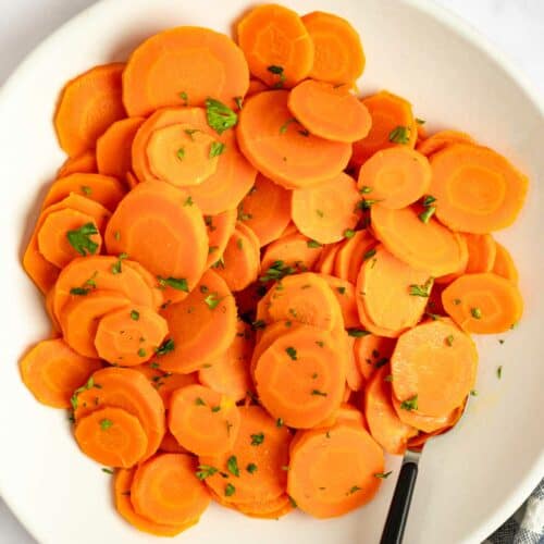 Cooked carrots on a plate and a bowl of chopped fresh parsley.