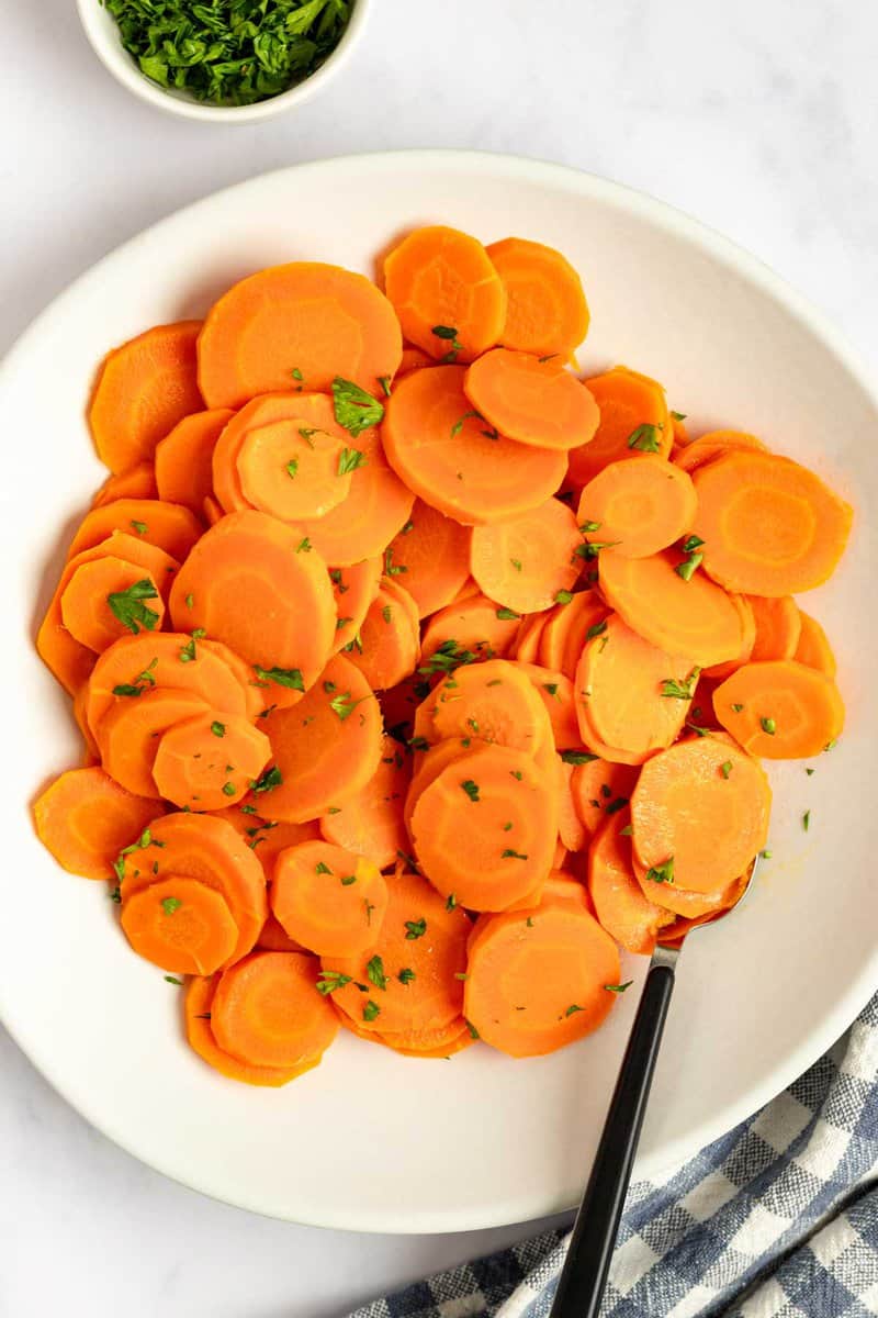 Cooked carrots on a plate and a bowl of chopped fresh parsley.