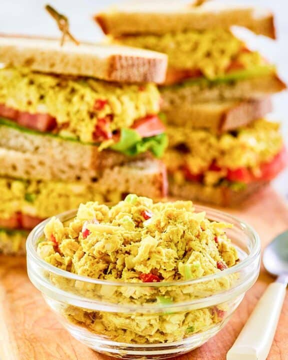 Curried chicken salad in a small glass bowl and sandwiches behind it.