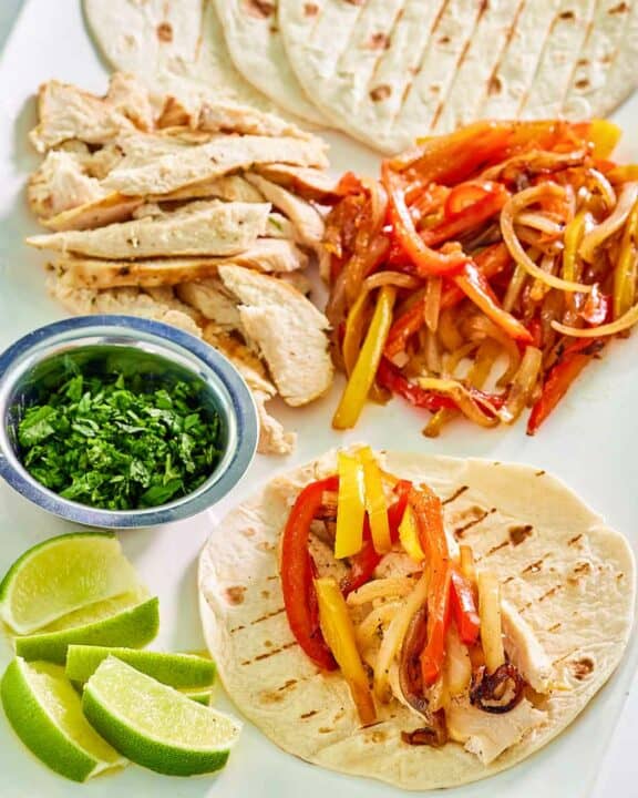 Grilled chicken fajitas, a cup of chopped cilantro, and lime wedges.