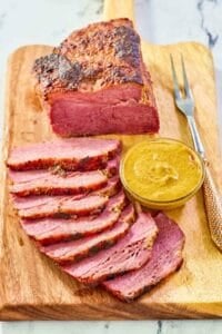 Baked corned beef, small bowl of mustard, and carving fork on a wood carving board.