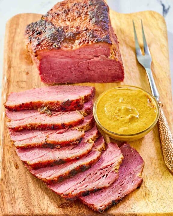 Baked corned beef, small bowl of mustard, and carving fork on a wood carving board.