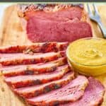Baked corned beef and a small bowl of mustard on a wood board.