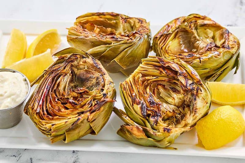 Grilled artichokes, a cup of garlic aioli, and lemon wedges on a platter.