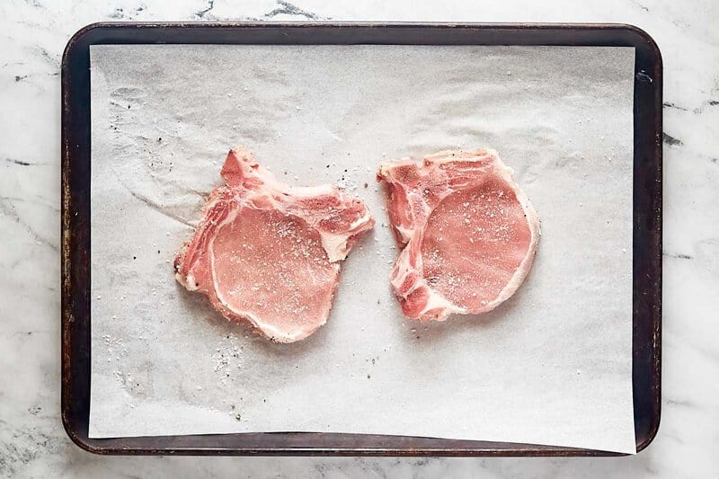 Two pork chops seasoned with brackish  and capsicum  connected  a parchment insubstantial  lined baking sheet.