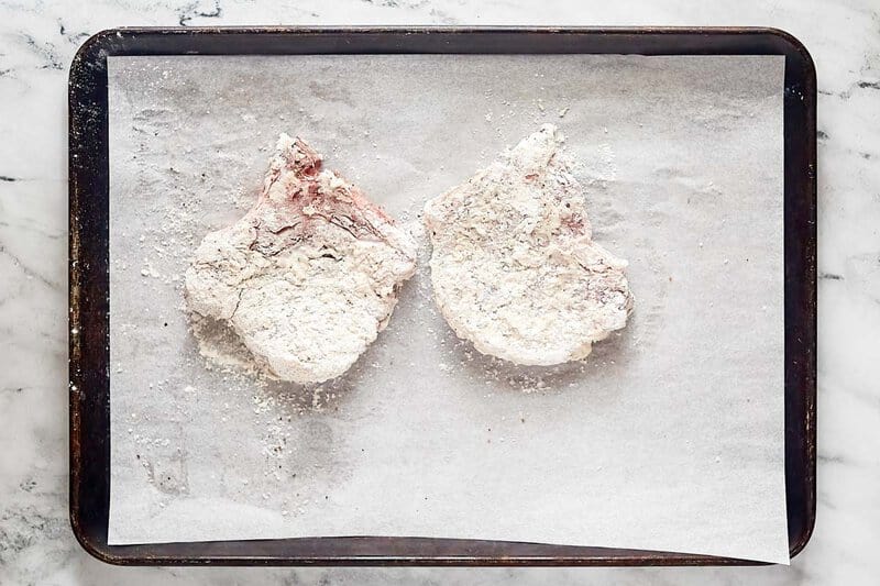 Two seasoned and breaded pork chops on a parchment paper lined baking sheet.