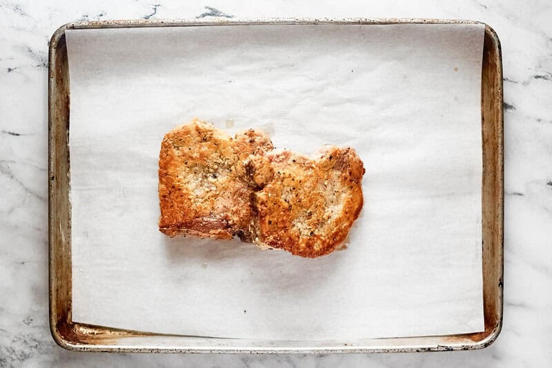 Two country fried pork chops on a parchment paper lined baking sheet.