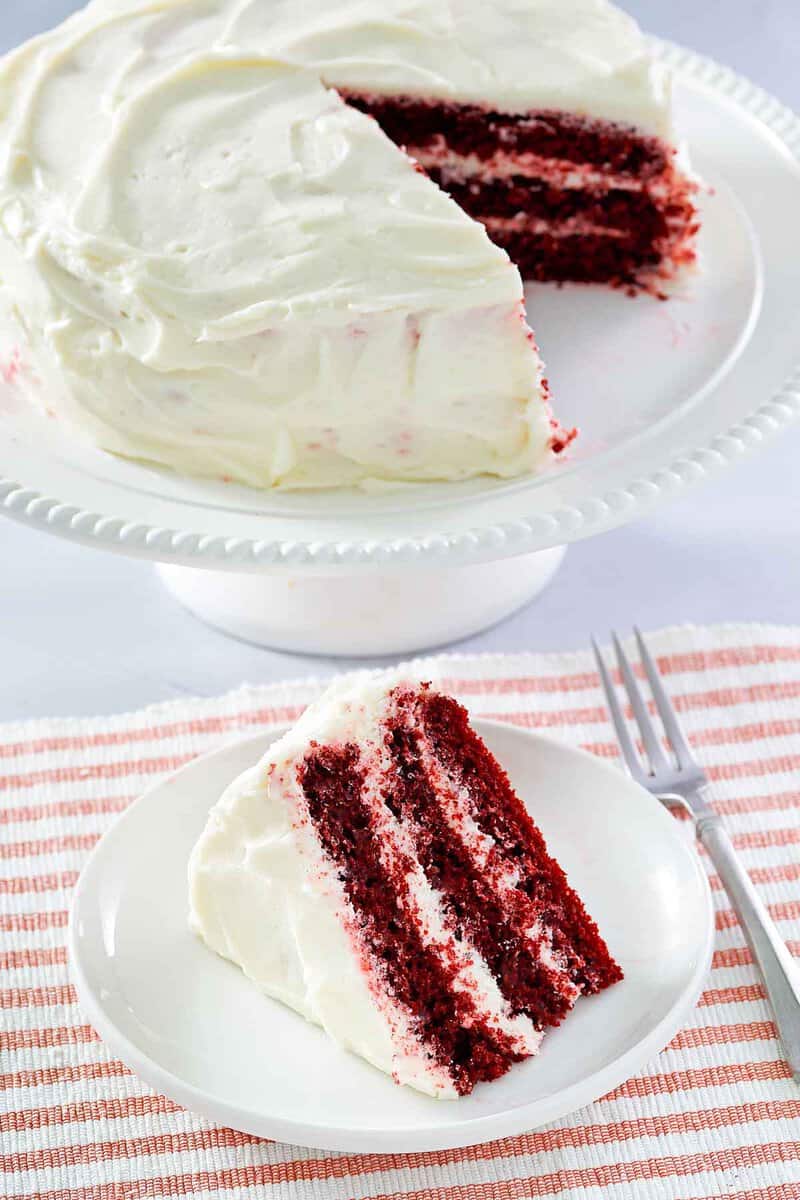 Copycat waldorf astoria red velvet cake slice on a plate and the cake behind it.